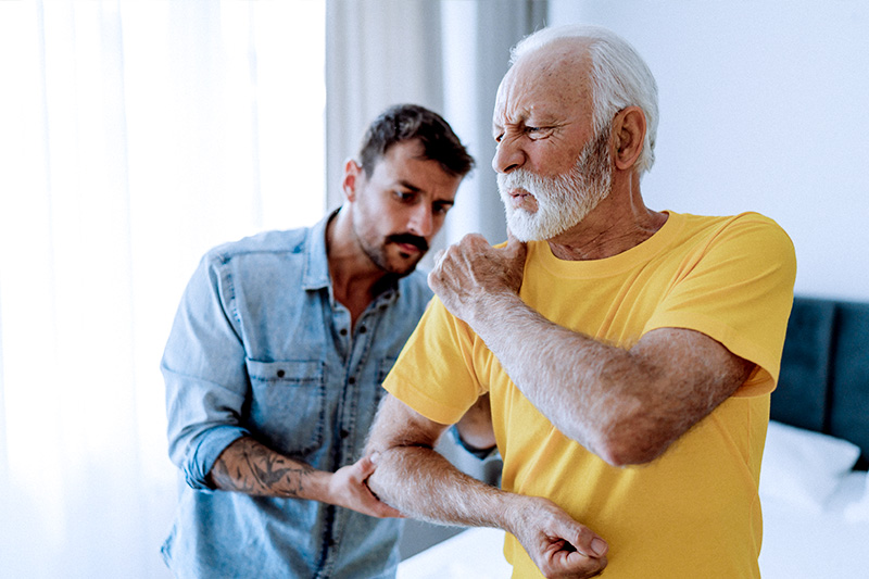Man with a chronic condition (arthritis) rubbing his shoulder while a person assists with his care.