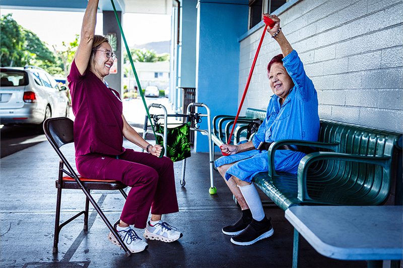 Cardiac occupational therapist and a woman sitting outside on benches, performing strength exercises and laughing together.